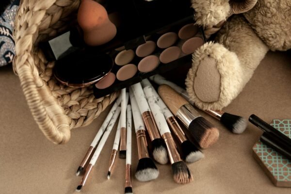 From above of various cosmetic products and makeup brushes in wicker basket placed near toy on beige table